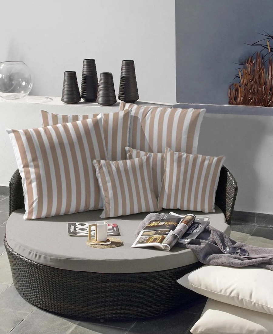 Reposa redonda para 2 personas. Es de ratán oscuro y sobre ella hay 6 cojines rayados blancos con beige.