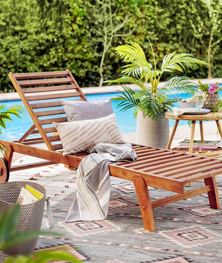 Reposera de madera junto a una piscina. Sobre ella hay 2 cojines y una manta en tonos neutros.