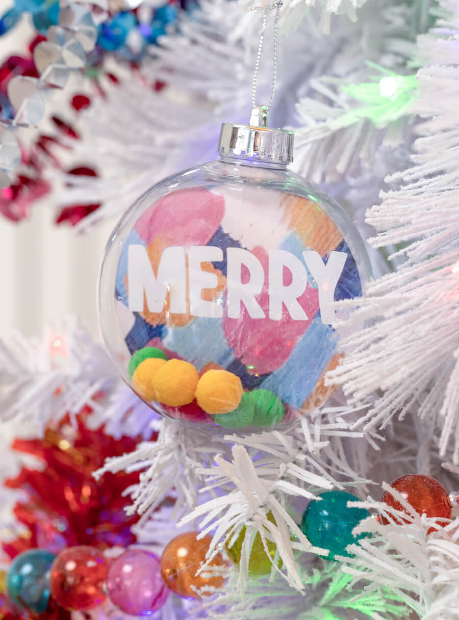 Detalle de ornamento navideño. Bola transparente con pompones de colores al interior. Al fondo tiene colores y la palabra "merry" escrita en blanco. Está colgado en un árbol de navidad blanco, junto a otros adornos de colores