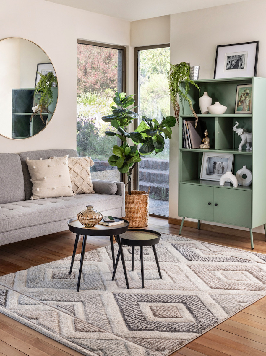 Living estilo nórdico. Repisa color matcha con adornos, libros y plantas. Sofá gris, set de mesas de centro redondas y alfombra gris con rombos. Gran espejo redondo