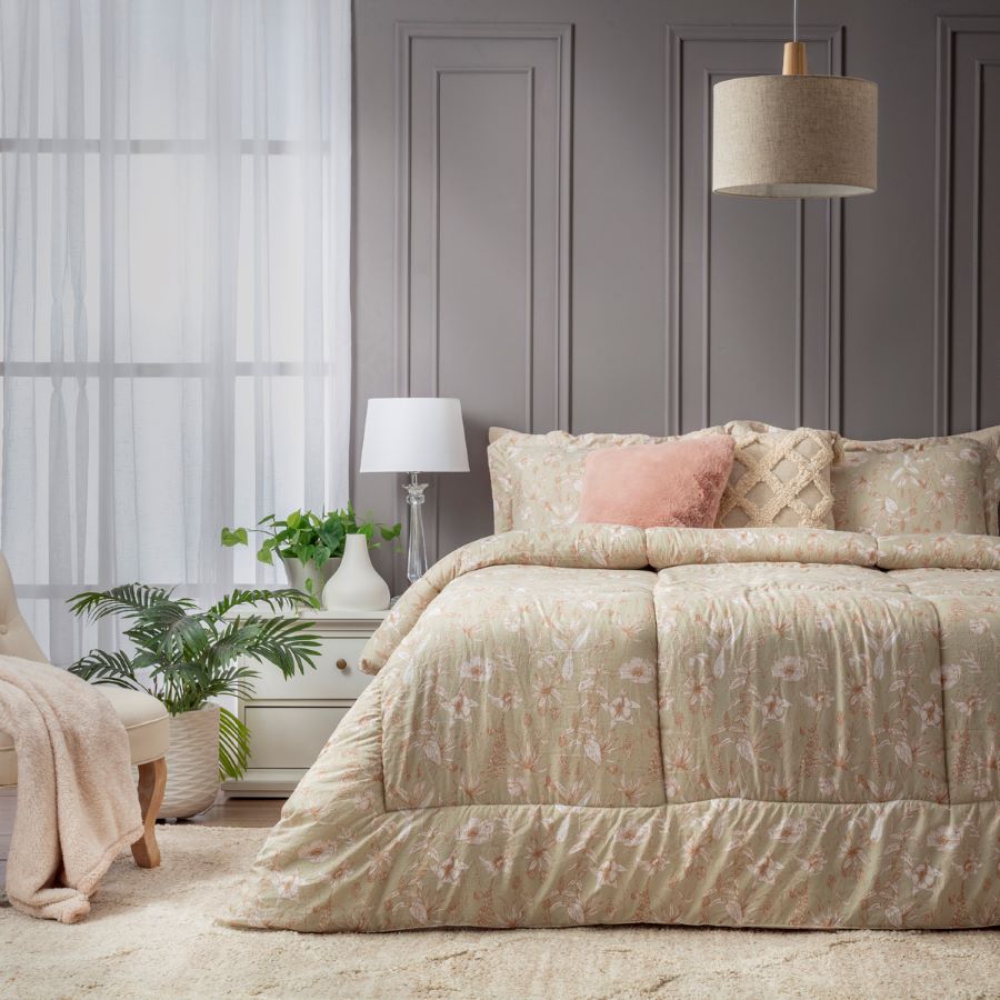 Dormitorio clásico. Cama con cojines en tonos pastel y beige, y el plumón tiene un patrón de flores. A la izquierda de la cama hay una mesita blanca con una lámpara de base transparente y algunas plantas decorativas.