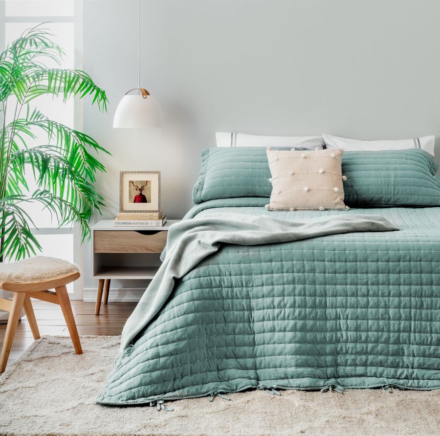 Dormitorio estilo nórdico con cama con ropa de cama en tonos verdes suaves. Cojines, uno de ellos beige con detalles de pompones. A la izquierda de la cama hay un velador de madera clara