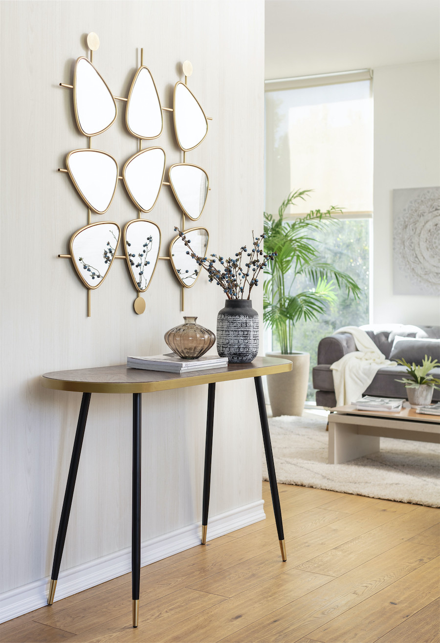 Recibidor con una mesa de arrimo con patas largas y delgadas negras, cubierta de madera oscura y bordes dorados. Sobre ella hay dos floreros y una pila de revistas. En la pared hay un espejo escultórico de 9 espejos más pequeños irregulares.