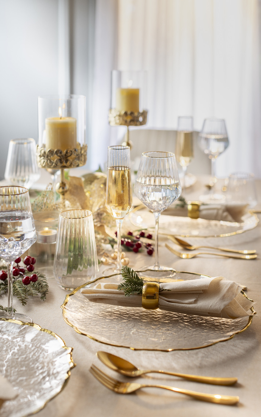 Mesa navideña elegante con copas doradas, platos con bordes dorados, velas y servilletas con aros festivos.