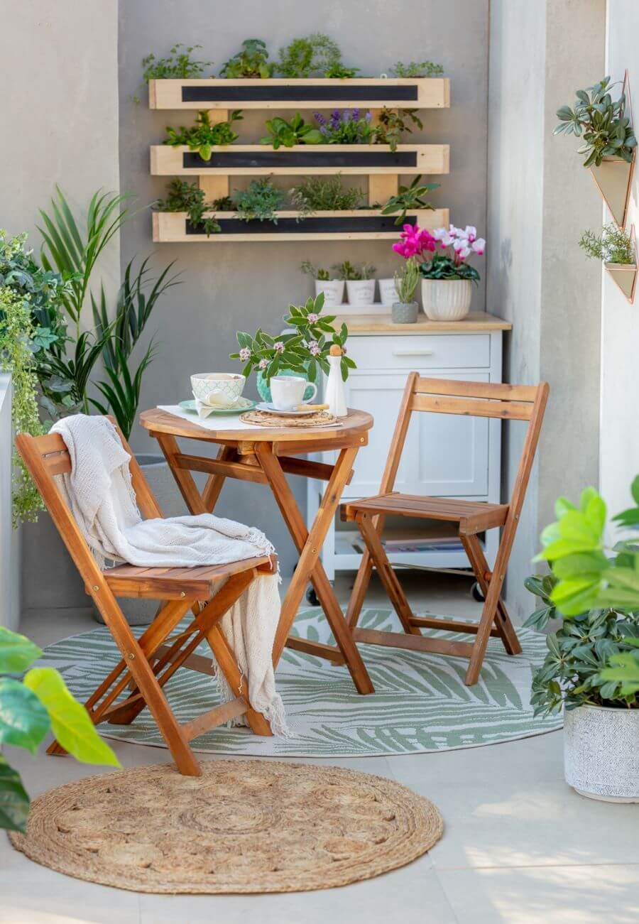Balcón con juego de terraza de madera, para dos personas, con mesa redonda y sillas plegables. Alfombra redonda con diseño de hoja tropical y una más pequeña, redonda, de yute. Mueble de madera con ruedas, repisas flotantes con plantas y hierbas