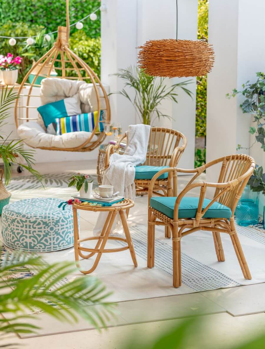 Juego de terraza de bambú. Mesa de centro pequeña, redonda y dos sitiales con cojines color turquesa. Silla colgante con cojines blancos y celestes. Gran alfombra de color blanco con líneas azules. Pouff redondo, de estilo boho de color celeste y blanco