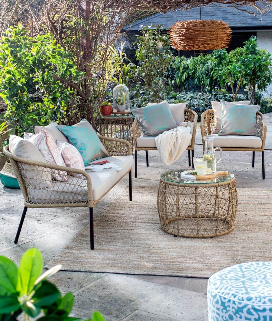 Terraza grande con juego de living compuesto por una mesa de centro redonda, un sofá y dos sillones hechos de ratán, con patas de fierro y cojines blancos. El suelo es de concreto y hay una gran alfombra color beige y café. Lámpara redonda de mimbre.