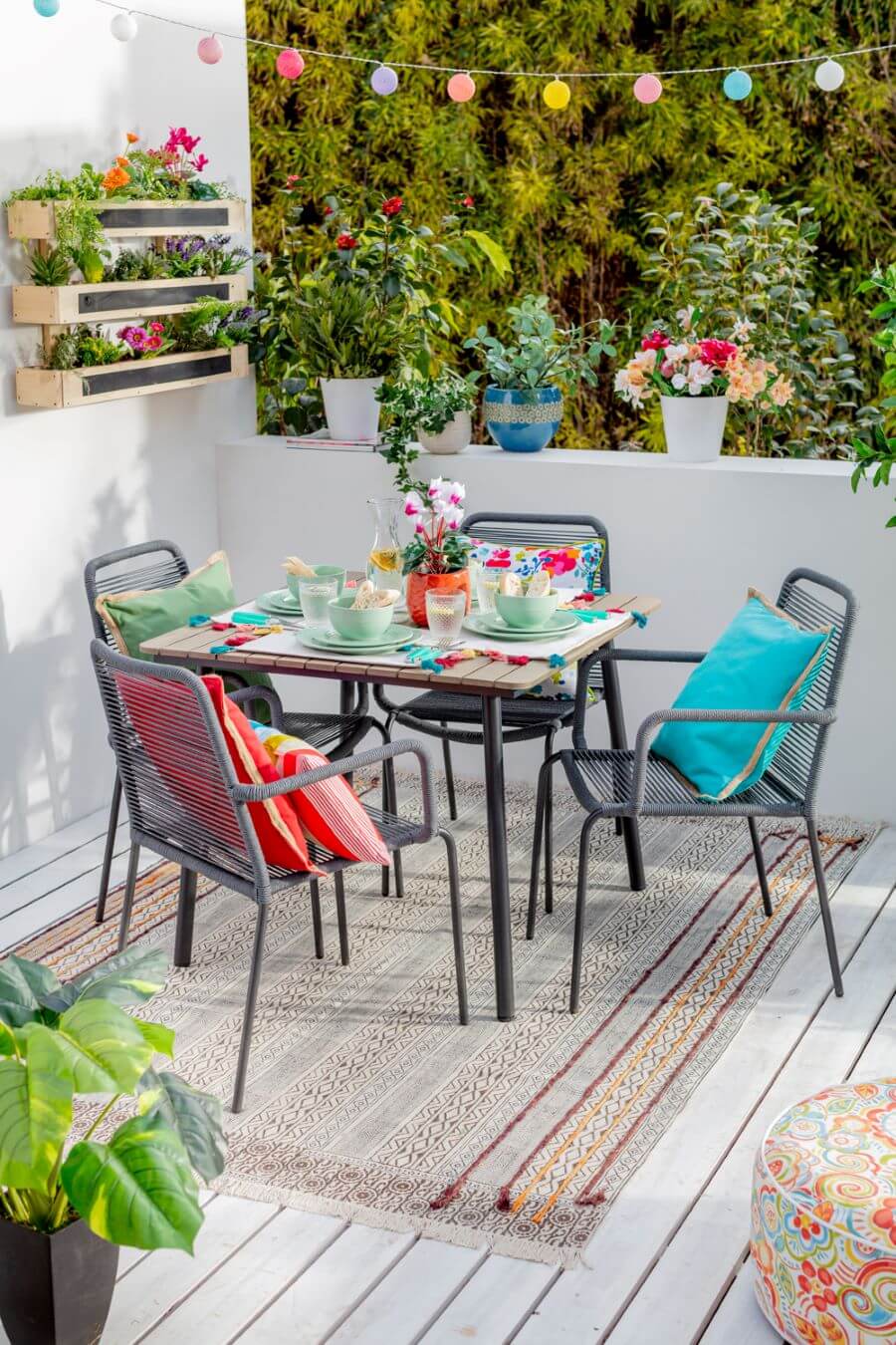 Terraza pequeña con mesa de metal cuadrada, con cuatro sillas. Mesa puesta con vajilla de colores. Alfombra grande con diseño boho sobre piso de madera blanco. Muro bajo con maceteros con plantas y flores. Repisas de muro de madera con plantas.