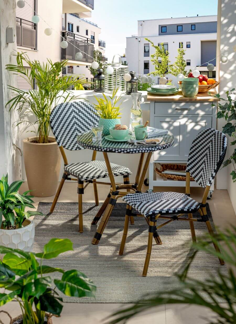 Balcón con juego de terraza para dos personas. Mesa redonda y sillas de estilo francés, de ratán blanco con negro. Alfombra color beige oscuro. Atrás hay una mesa con ruedas blanca, de madera, con vajilla encima. En una esquina hay una gran planta