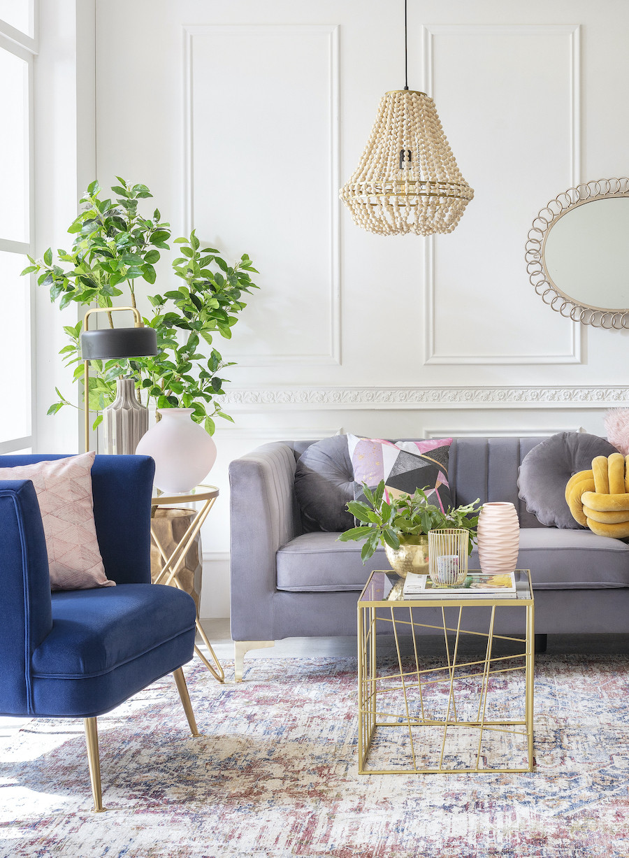 Sala de estar de estilo clásico chic con paredes blancas con molduras, una alfombra de colores, un sofá gris oscuro, una poltrona azul marino. También hay una pequeña mesa de centro dorada con adornos sobre ella. Al costado del sofá hay plantas.