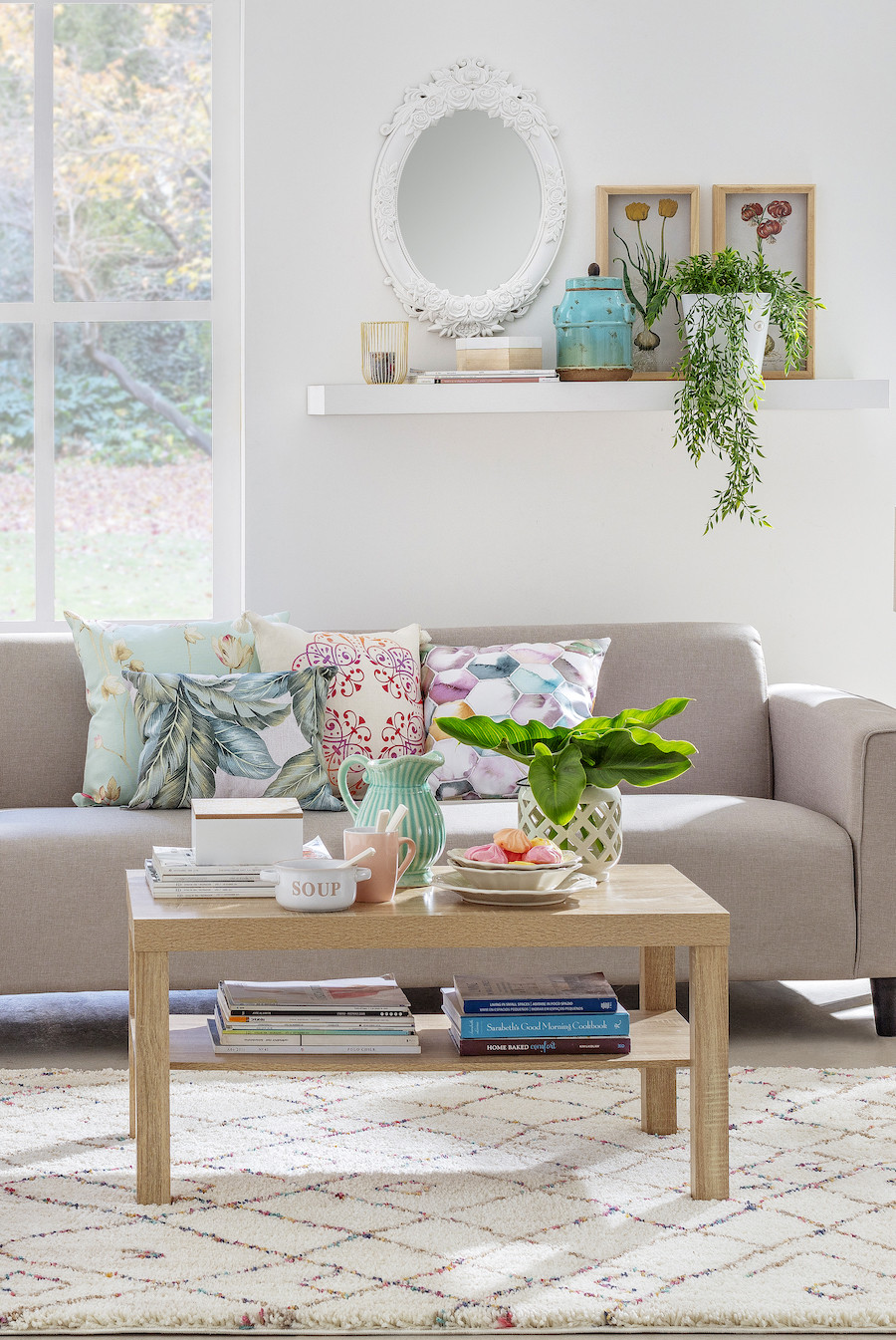 Living de tonos neutros con una pared blanca, sofá beige y cojines de colores. Al frente hay una mesa de centro de madera con plantas, libros y adornos sobre ella.