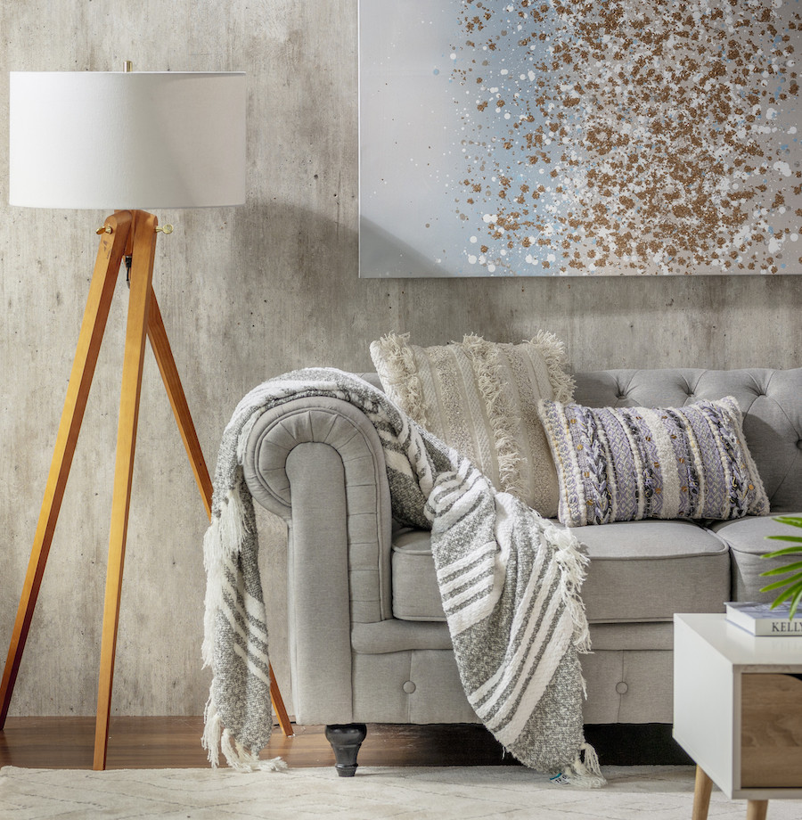 Detalle de un living con un sofá gris claro, cojines de tonos neutros y una manta rayada blanca con gris. Al costado hay una lámpara de pie de madera con pantalla blanca.