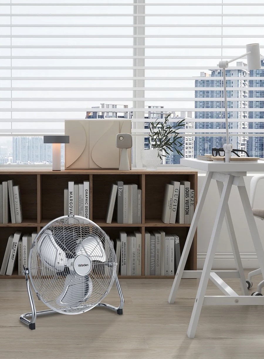 Home office con un estante bajo con libros y revistas, un escritorio blanco y un ventilador metálico a nivel del piso.