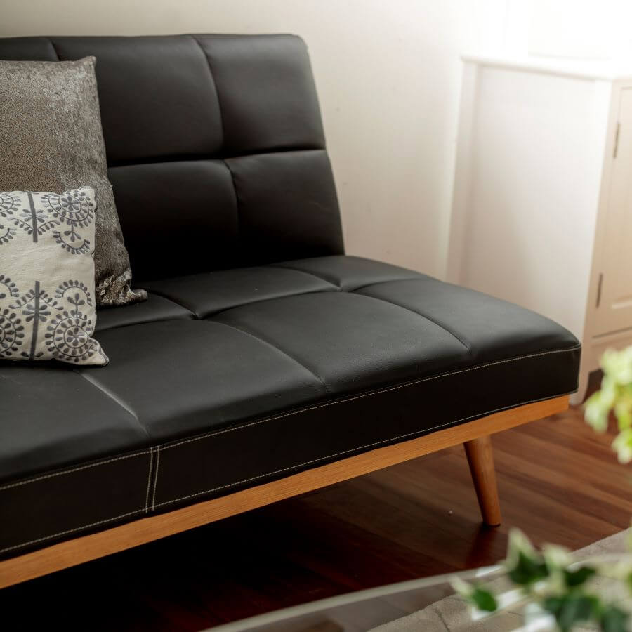 Detalle de sillón o futón de ecocuero negro, con patas de madera, Tiene dos cojines, uno metálico gris y otro blanco con bordados azules. Piso de madera oscura y muro blanco.