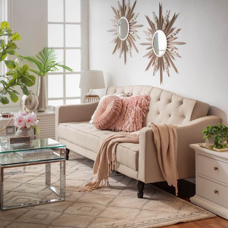 Living de estilo clásico, con sillón de tela color beige, con respaldo capitoné. Cojines y manta rosada. Mesa de centro de vidrio con patas plateadas. En el muro blanco hay dos espejos redondos con marco tipo estrella. Alfombra beige con líneas grises.