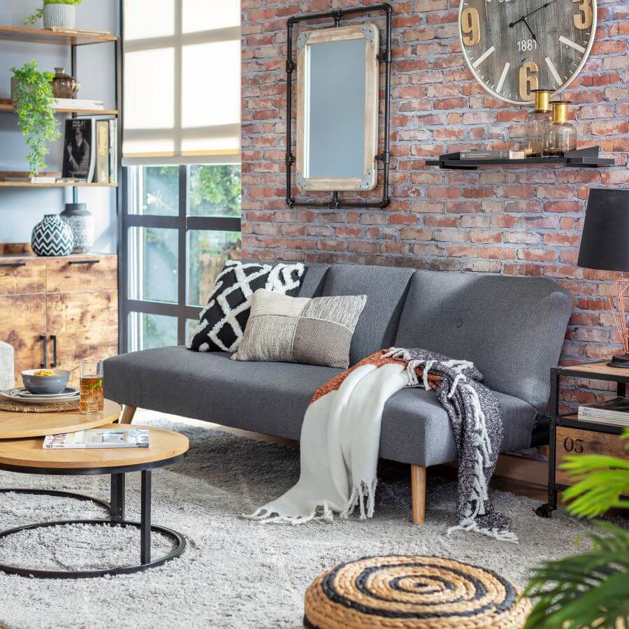 Living industrial con muros de ladrillo. Sillón de tela gris y patas de madera, con cojines y una manta encima. Mesa de centro doble, redonda, de base de madera y patas de fierro. Gran alfombra de color gris. Repisa de madera con fierro.