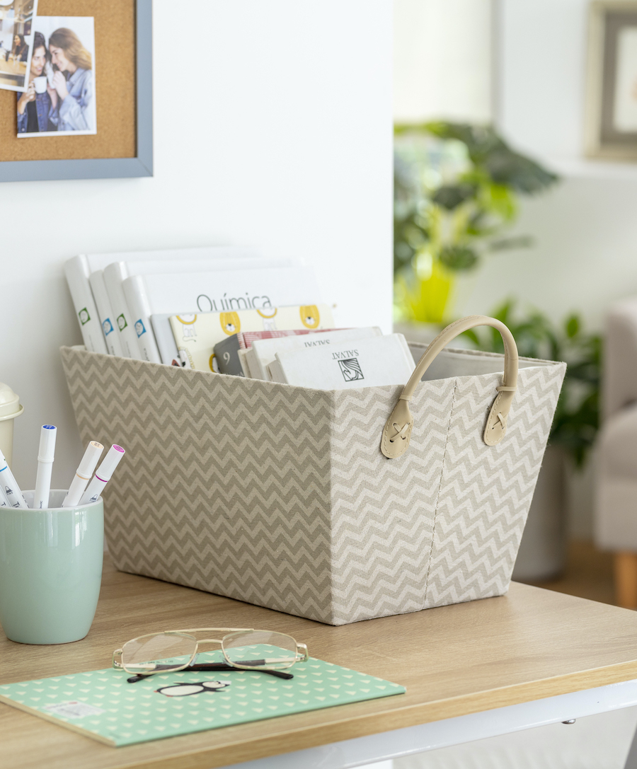 Caja organizadora de tela con diseño zigzag en color beige. En su interior hay libros y libretas. Al costado hay una taza con lápices y unos anteojos.