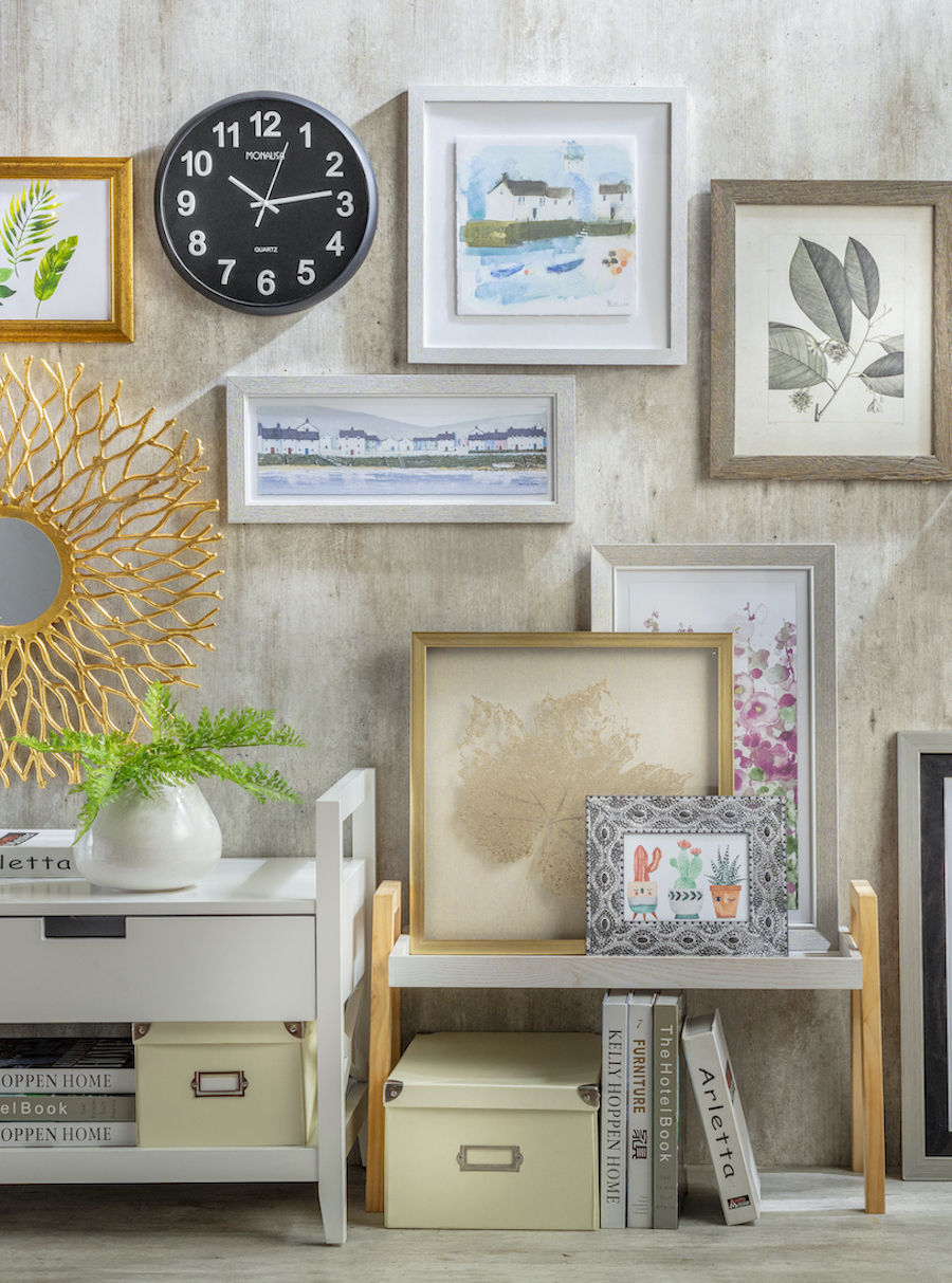 Pared de una sala de estar con diferentes cuadros con ilustraciones de hojas en tonos neutros y pastel, un espejo negro y un reloj con marco dorado con forma de sol.