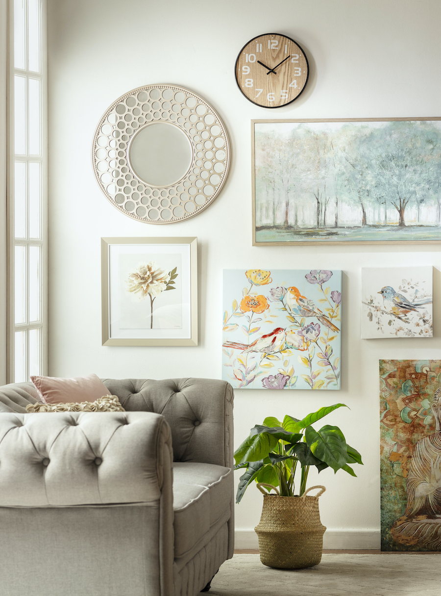 Rincón de una sala de estar con un sofá capitoné gris con cojines rosa pastel, una planta artificial dentro de un canasto y un muro galería en colores pastel en la pared del fondo.