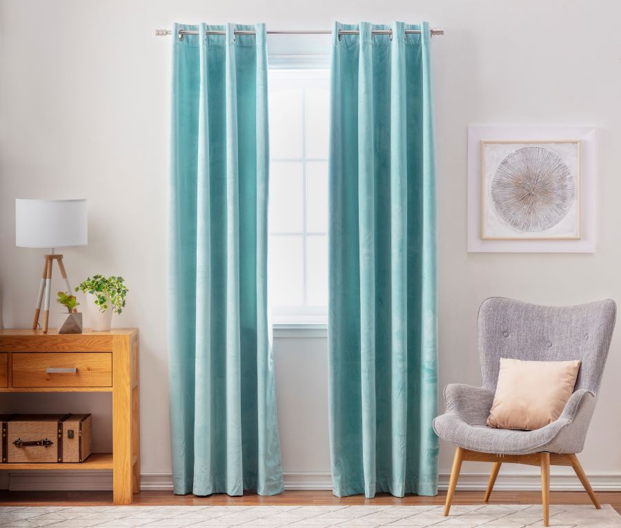 Living clásico con ventana central, con cortinas de tela color turquesa que llegan al piso. Al lado hay una poltrona gris y patas de madera con un cojín beige, y en el muro un cuadro con un dibujo de flor. Al otro costado hay un arrimo de madera