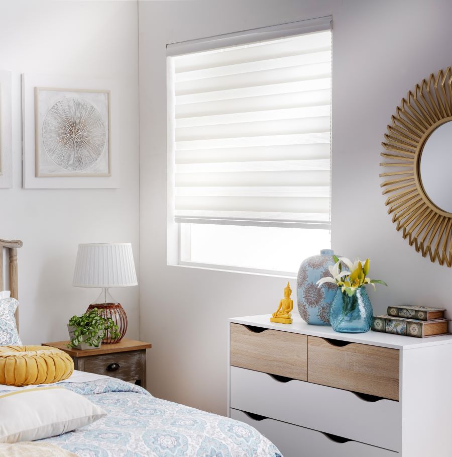 Dormitorio con ventana con una cortina roller duo color blanco. Bajo la ventana hay una cajonera estilo nórdico, con cajones y blancos y de madera. Sobre ella hay adornos y en la pared un espejo redondo. Cama de dos plazas y junto a ella un velador