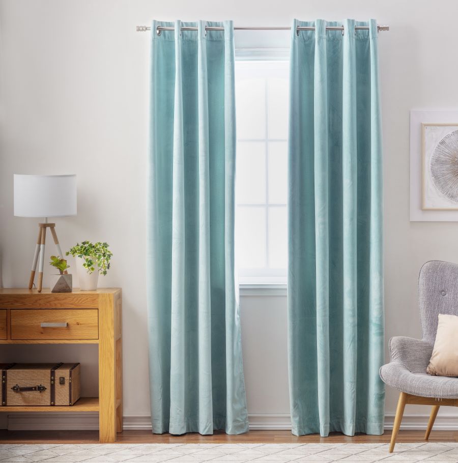 Ventana con una cortina de tela color celeste o turquesa. Junto a ella hay un arrimo de madera, con un cajón y en la parte inferior una repisa. Sobre él hay una lámpara con tres patas de madera y pantalla redonda blanca. Al otro lado hay un sitial gris