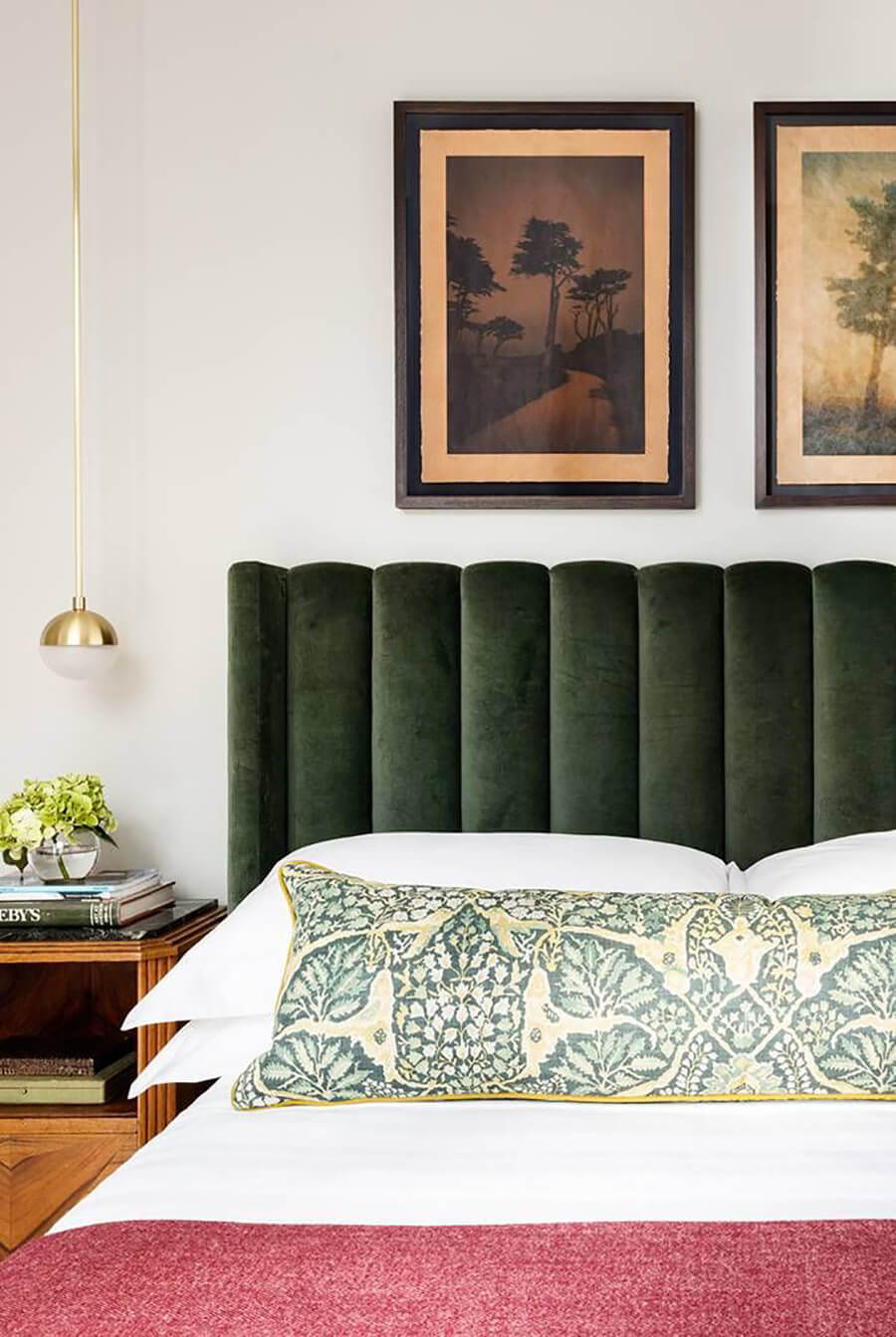 Dormitorio de estilo clásico con una cama con respaldo velvet verde, sábanas blancas, piecera burdeos y cojín alargado con estampado verde y amarillo. El velador a su costado es de madera y sobre él cuelga una lámpara dorada en forma de esfera. En la pared hay dos cuadros de árboles con marcos de madera oscura.