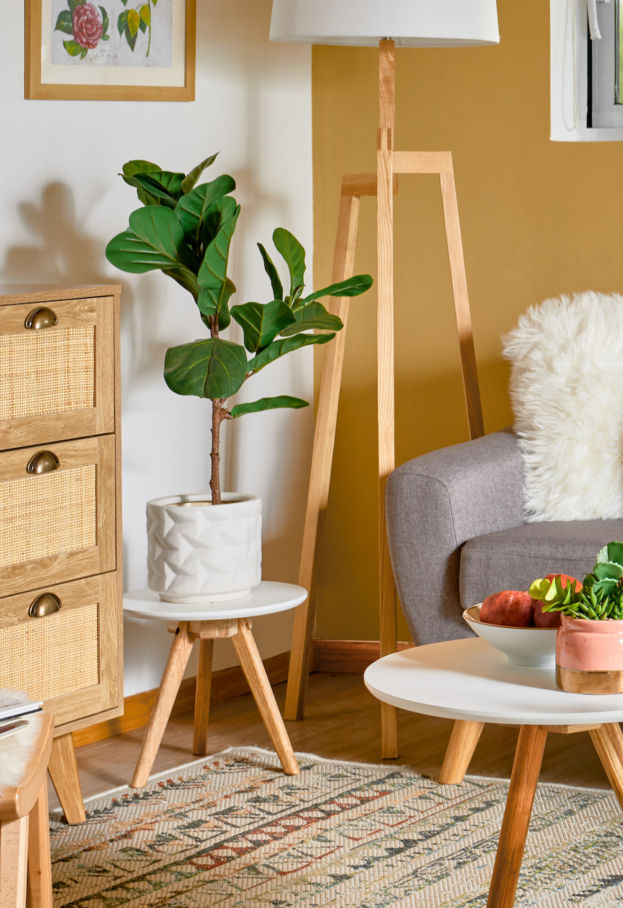 Detalle de una sala de estar de paredes blancas y mostaza con un sofá gris, lámpara de pie de estilo nórdico y junto a ella, una mesa auxiliar de patas de madera y superficie blanca. Sobre ella hay un macetero blanco con una planta artificial ficus lirata.