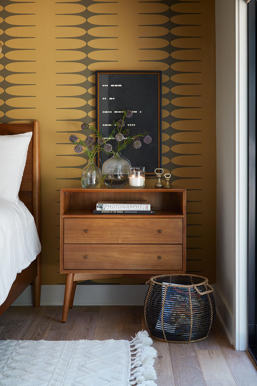 Velador de madera oscura con estilo vintage y patas cónicas. A su lado hay un canasto de fibras naturales, una alfombra blanca con diseño y al otro costado, parte de una cama de madera con ropa de cama blanca. La pared del fondo tiene un papel mural retro.