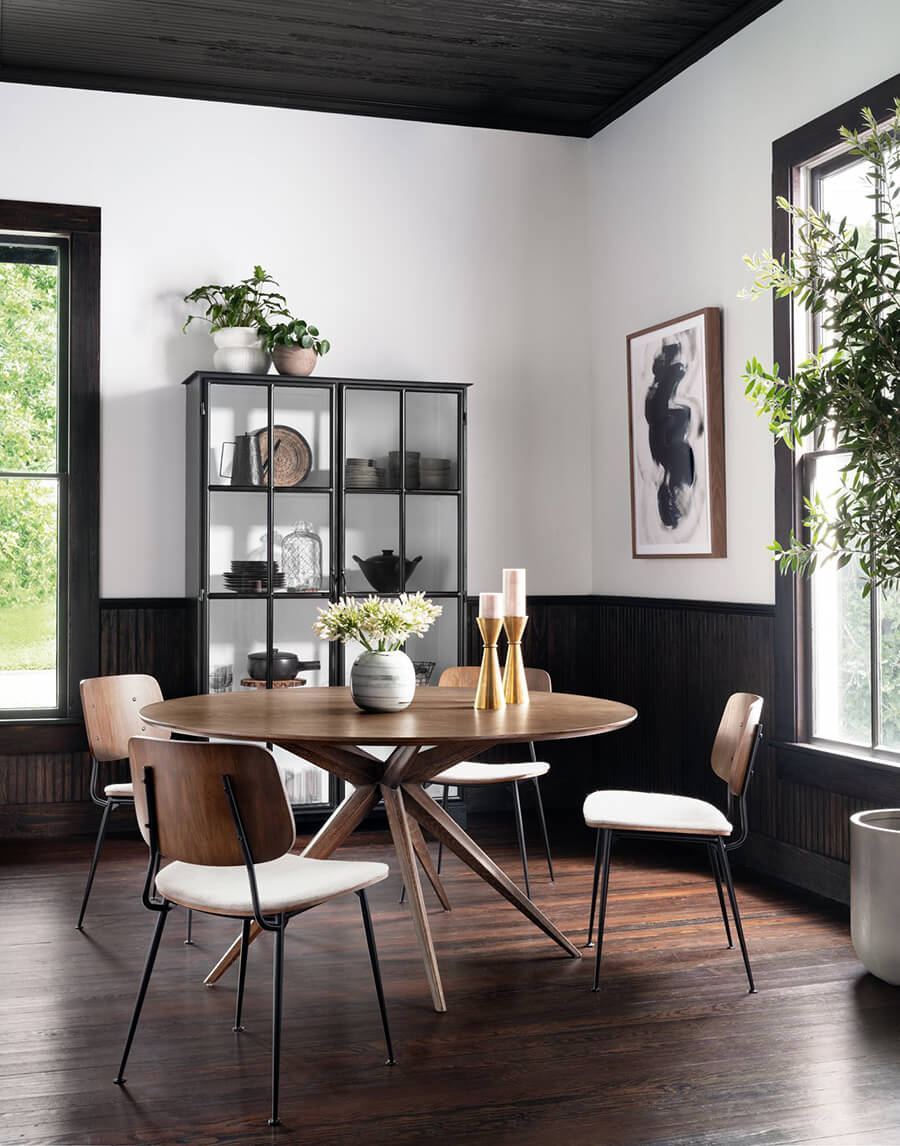 Juego de comedor de madera. La mesa es circular y las sillas tienen un look vintage con formas redondeadas en su respaldo y asiento. Hacia el fondo hay una vitrina negra con loza y menaje en su interior. Se ubicada junto a una venta con marco negro. En la otra pared también hay una ventana, junto a un cuadro con una ilustración y una gran planta de interior.