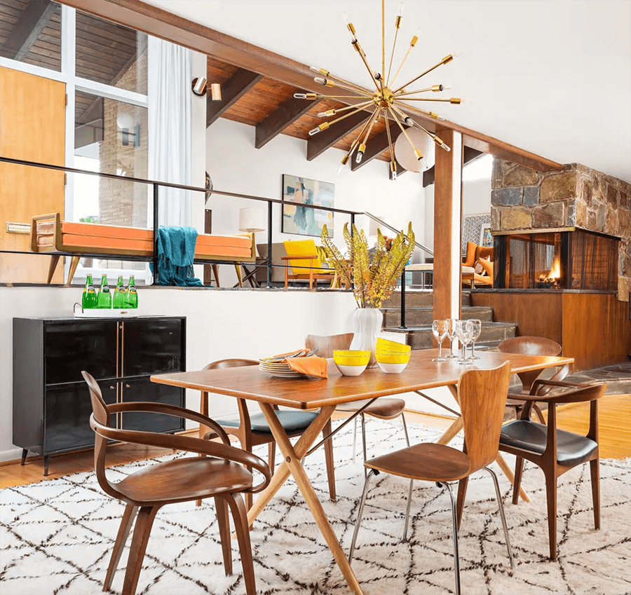 Espacio de estilo vintage con comedor de madera y sillas, también de madera, pero en diferente estilo. Desde el techo cuelga una lámpara escultórica con acabado dorado y ampolletas decorativas. El suelo es de madera y hay una gran alfombra blanca con líneas café.