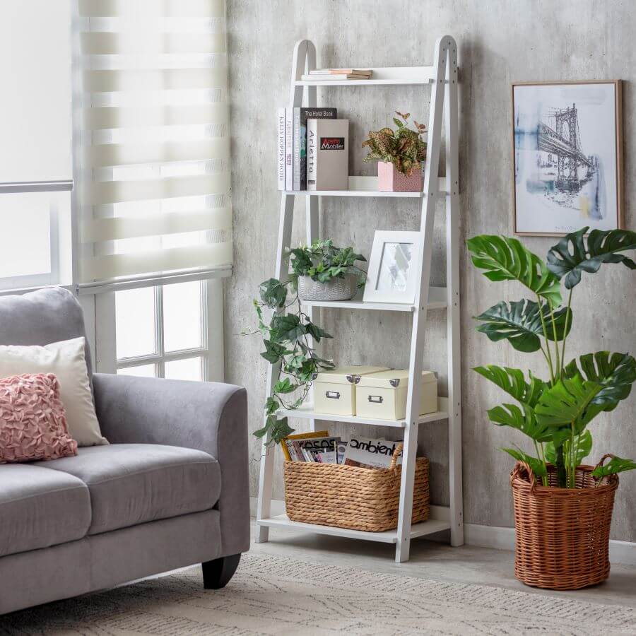 Living comedor pequeño, con una repisa tipo escalera, de madera y color blanco. Tiene 5 niveles en los que hay libros, plantas, cuadros y un canasto de mimbre. Gran planta en un macetero de mimbre y un sofá color gris con dos cojines.