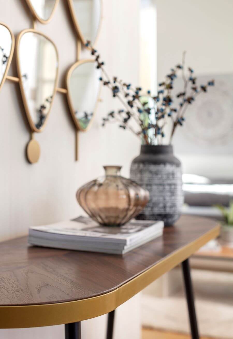 Arrimo de madera oscura, ovalado. Sobre él hay revistas, un florero de vidrio y un gran jarrón de cerámica con ramas. En el muro blanco hay un espejo con varias secciones, estilo mid century, con marco dorado.