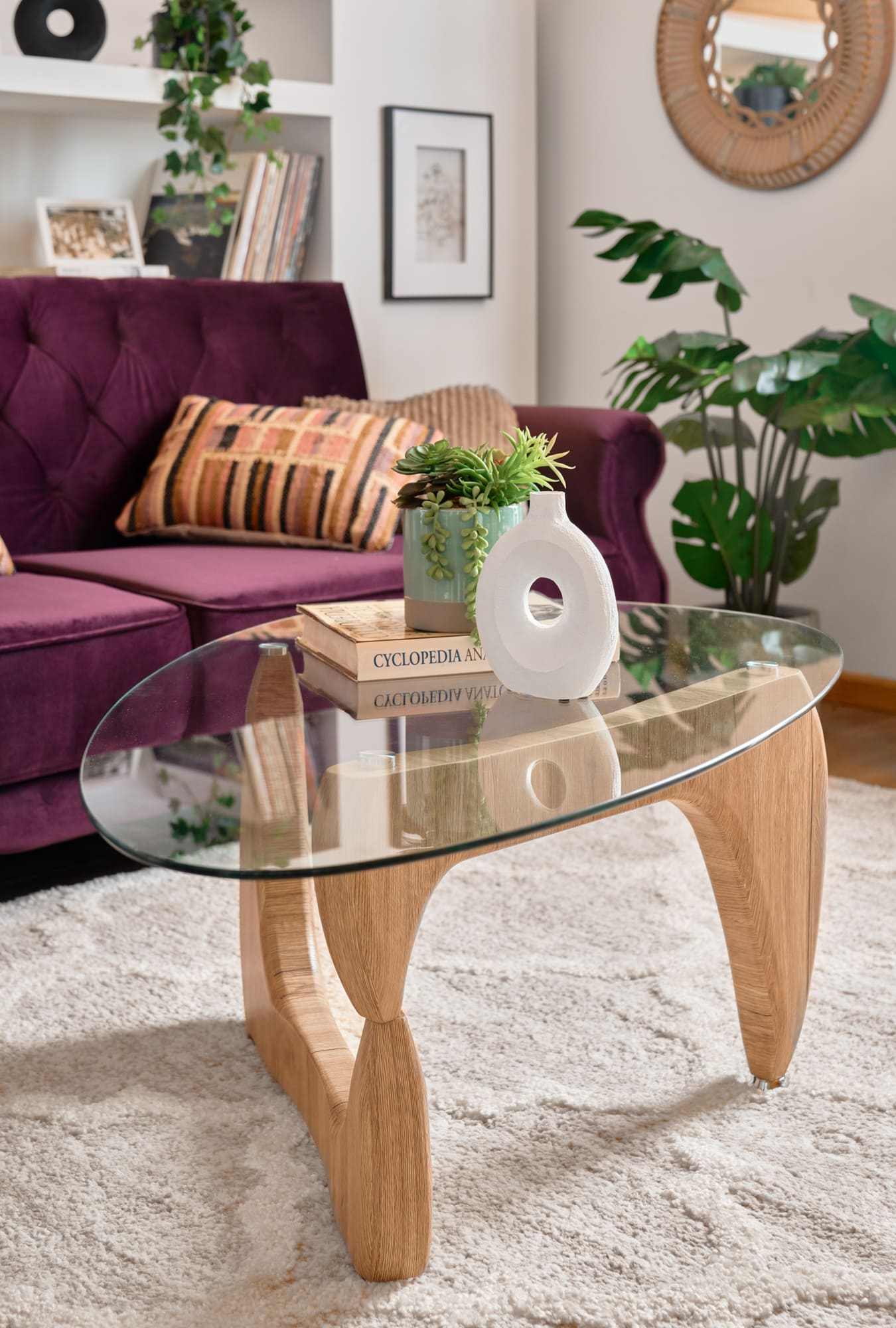 Mesa de centro de vidrio ovalado con patas de madera. Sobre la mesa hay una maceta con plantas sobre un libro y un jarrón decorativo blanco. Al fondo hay un futón morado con respaldo estilo capitoné y una planta.