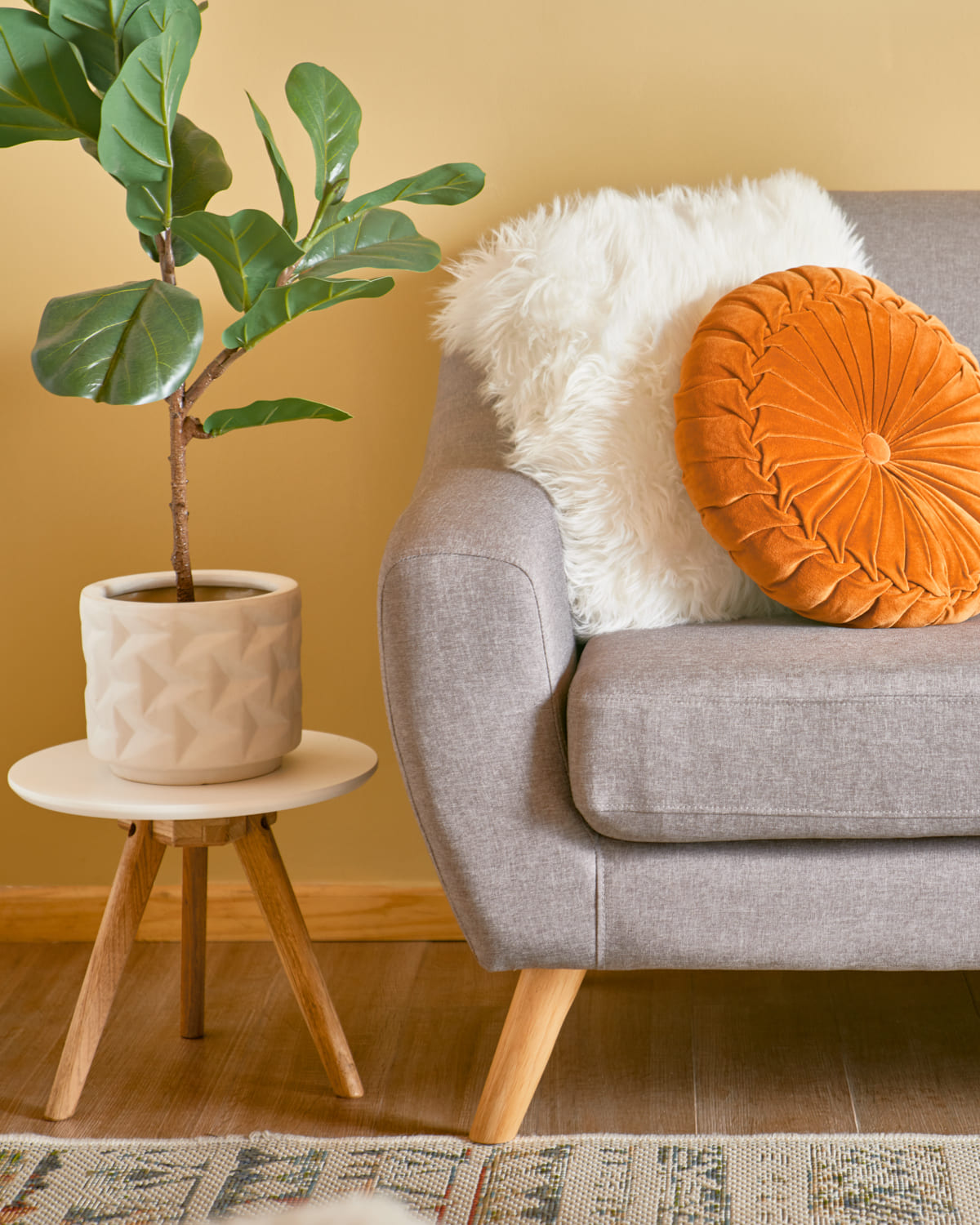 Sofá gris con patas de madera y una mesa lateral blanca y redonda con patas de madera y que sostiene una maceta blanca con una planta. Están frente a una pared amarilla.