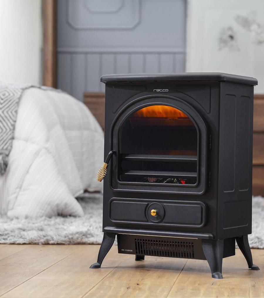 Estufa eléctrica con diseño vintage, de color negro, disponible en Sodimac. Es de metal y está ubicada sobre un piso de madera clara. Atrás se ve una cama con plumón blanco y una manta.