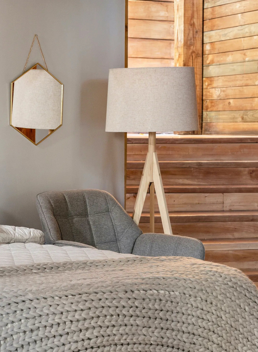 Detalle de un dormitorio en tonos grises con una cama con una piecera gris clara, una poltrona gris oscura y una lámpara de pie de estructura de madera y pantalla de tela blanca.