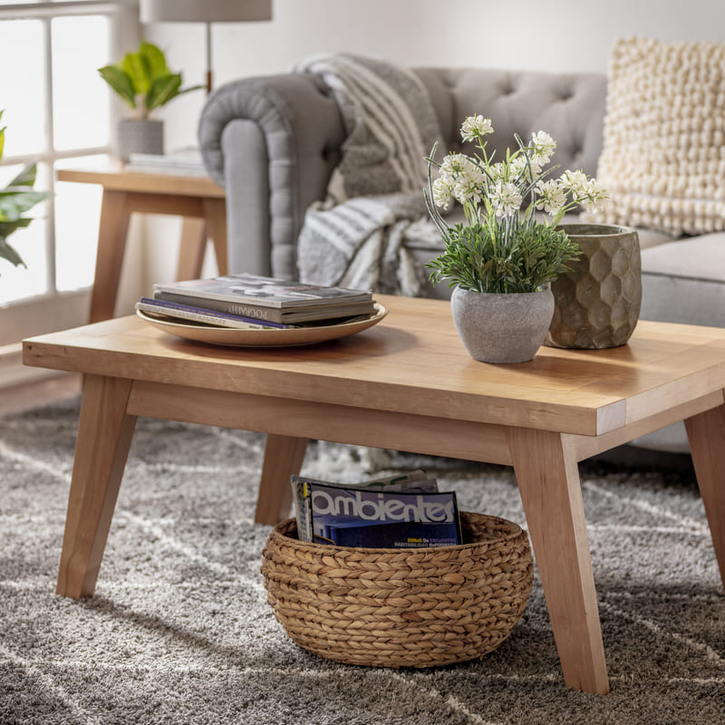 Mesa de centro rectangular de madera clara. Sobre la mesa hay decoraciones como macetas y bandeja y, debajo, hay una canasta de mimbre con revistas.