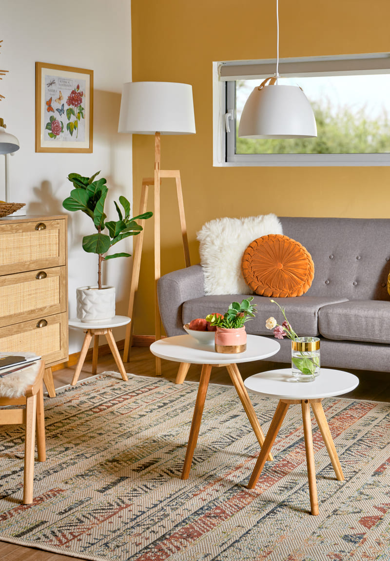 Living con un sofá de dos cuerpos gris y tres mesas similares: dos funcionan como mesa de centro y una como mesa lateral. Tienen la cubierta blanca y patas cónicas de madera clara.