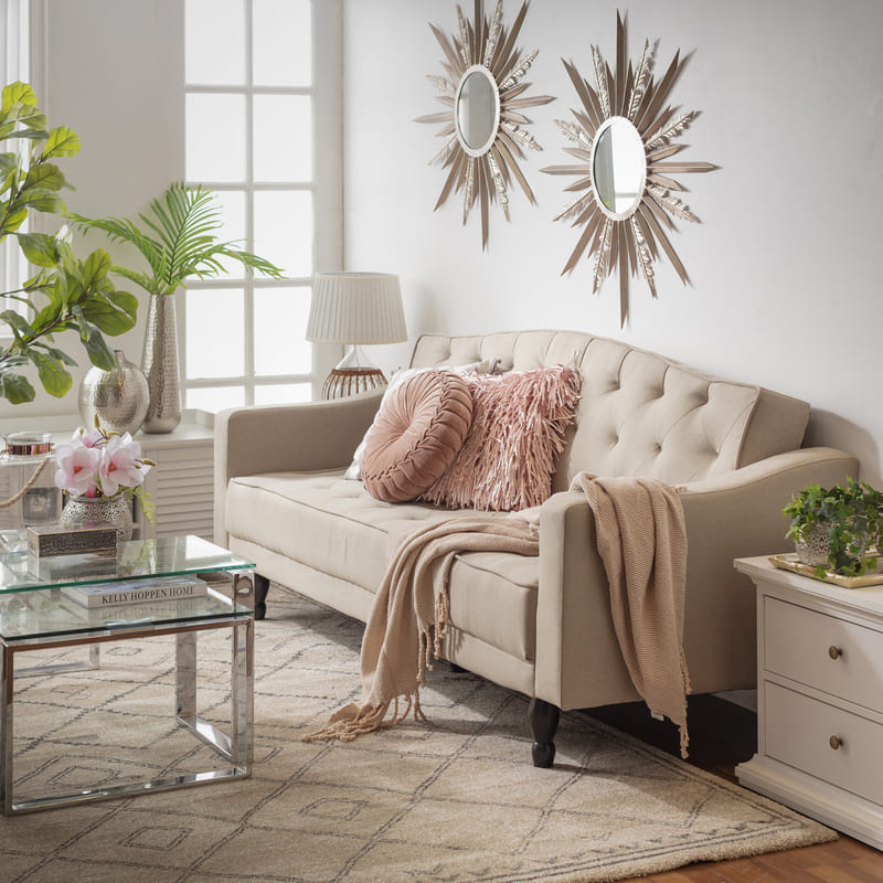 Sala de estar con un sofá de tres cuerpos beige con respaldo estilo capitoné, sobre el sofá hay dos cojines rosa y una manta rosa palo. El sofá está sobre una alfombra blanca shaggy que también contiene una mesa de centro rectangular de vidrio.