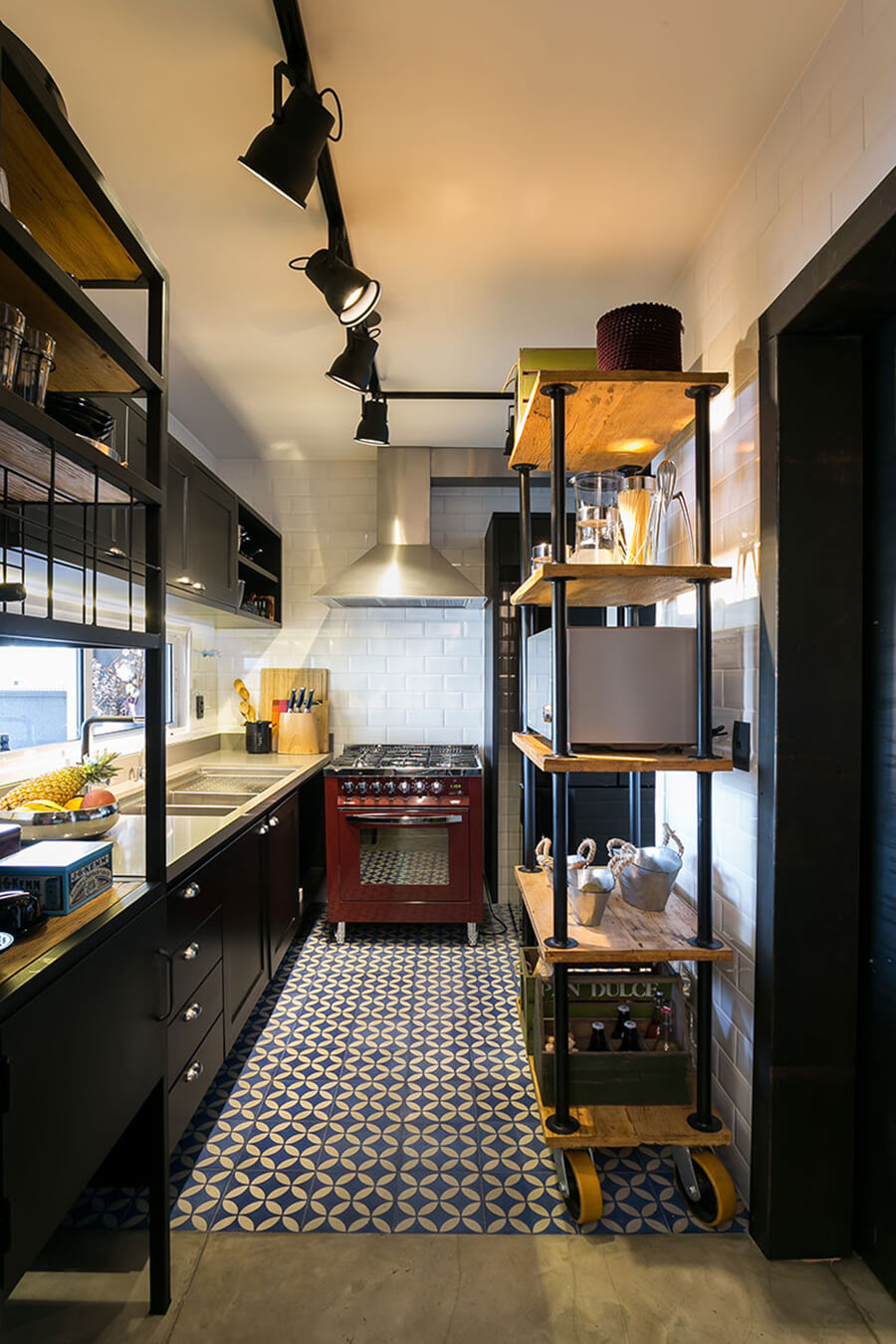 Cocina de estilo industrial con dos estantes de estructura metálica negra y superficies de madera. En el techo hay una lámpara que une cuatro fotos que iluminan el espacio. Los muebles de cocina son negro con cubierta blanca y la cocina es roja. Mientras que la campana sobre ella es metálica.