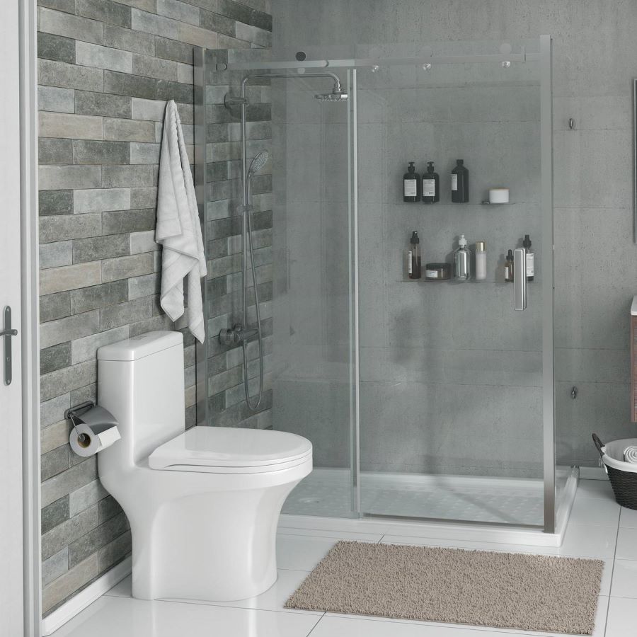 Baño moderno con inodoro de loza blanco. Muros con cerámicos grises, simil concreto, alargados y de diferentes tonalidades. Ducha con mampara de vidrio y dos repisas flotantes con artículos de aseo. Piso de cerámicos blancos.