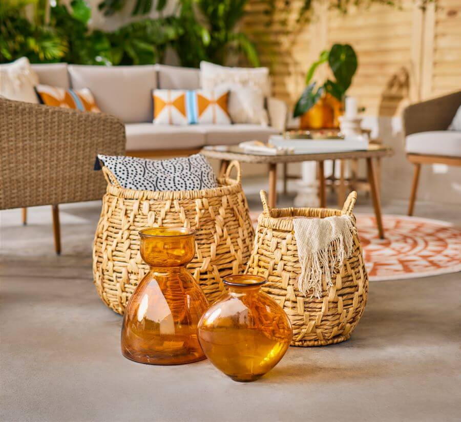 Detalle de dos canastos de mimbre. El más grande tiene un cojín adentro y el otro una manta color beige. Frente a los canastos hay dos jarrones o botellas de vidrio color ámbar. Atrás se de una terraza amplia, con un juego de living
