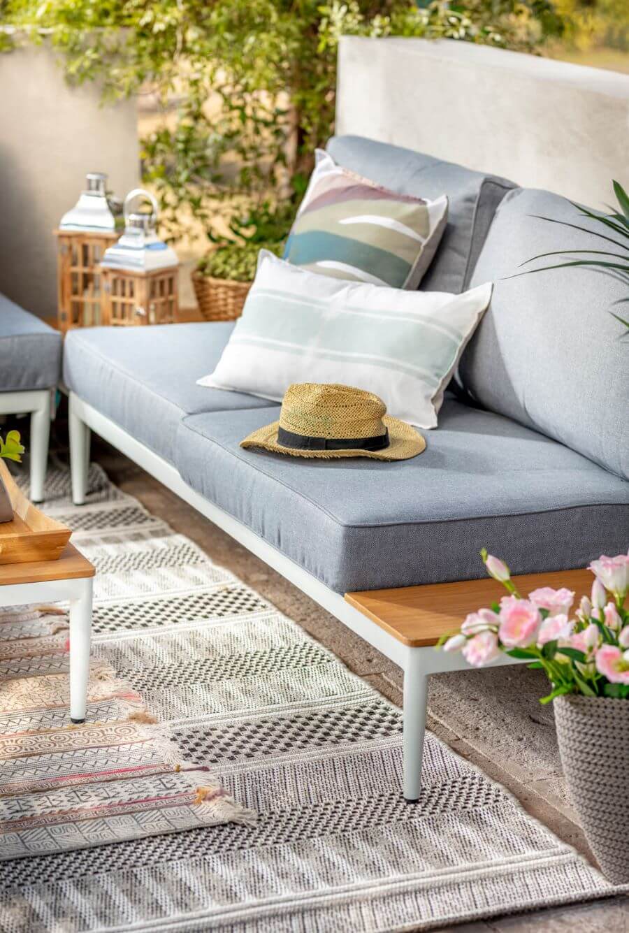 Juego de living de terraza. Detalle en el sillón de dos cuerpos, de base metálica blanca con cojines grises. Tiene incorporada una mesa de madera al costado. Sobre el sillón hay dos cojines y un sombrero. 
