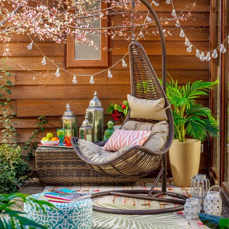 Balcón con muro de revestimiento de madera rojiza. Hay una silla colgante con cojines. Frente a ella, en el suelo, hay farones con velas y un pouff celeste. Atrás hay una banqueta de mimbre, con más faroles, y una planta en un macetero color mostaza
