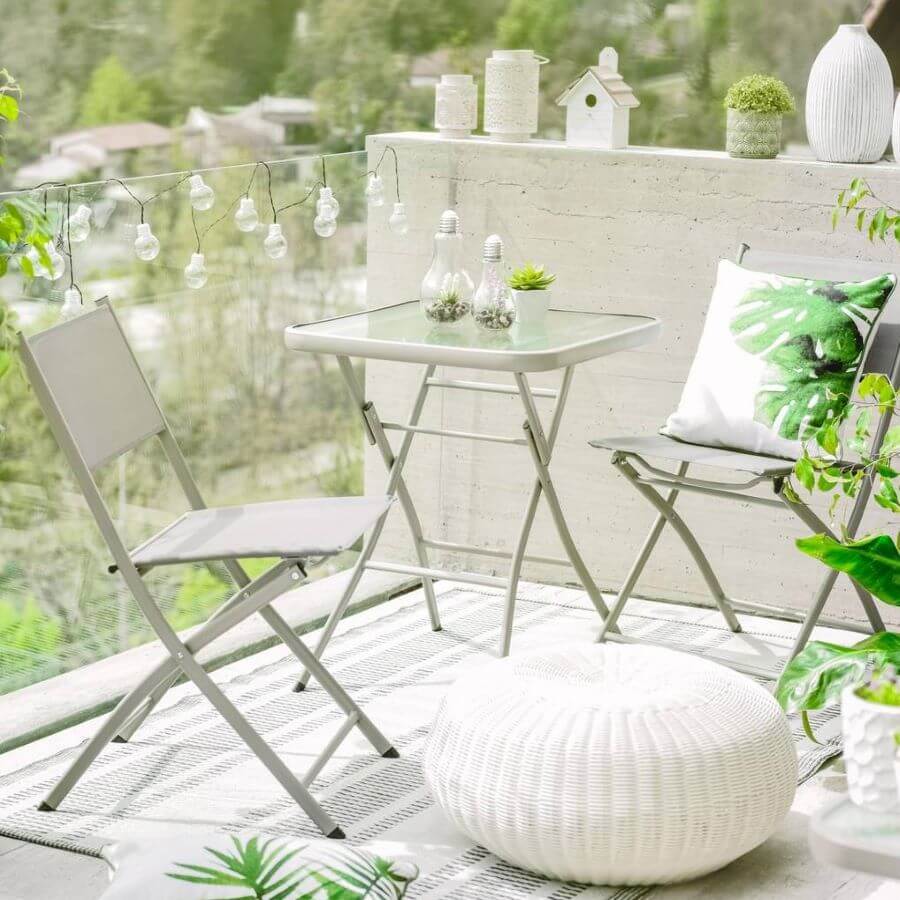 Terraza pequeña, moderna y blanca.  Juego de balcón para dos personas, metálico plegable. La mesa es cuadrada con base de vidrio. En el piso hay una alfombra blanco con gris y un pouff tejido blanco. Hay guirnalda de luces en la baranda
