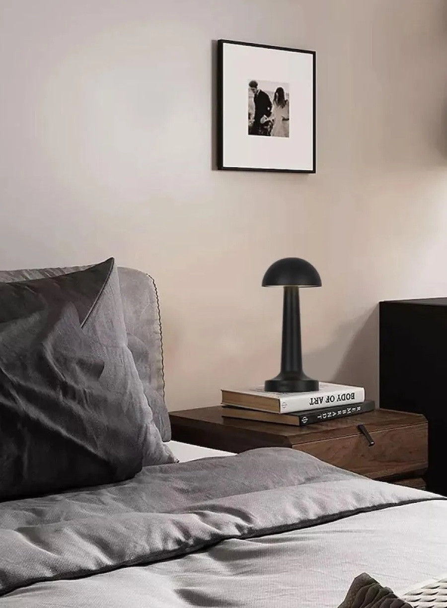 Dormitorio de estilo Ejecutivo Urbano con una cama con sábanas grises, un velador de madera oscura y sobre él, dos libros y una lámpara inalámbrica negra mate con forma de hongo.