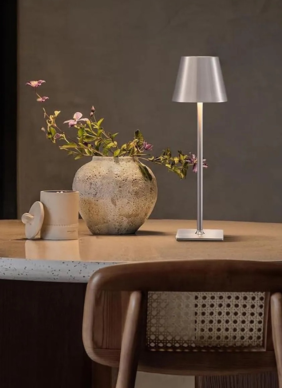 Detalle de una mesa de comedor con una lámpara inalámbrica plateada que entrega un punto de luz cálido, un jarrón con flores y un frasco color beige.
