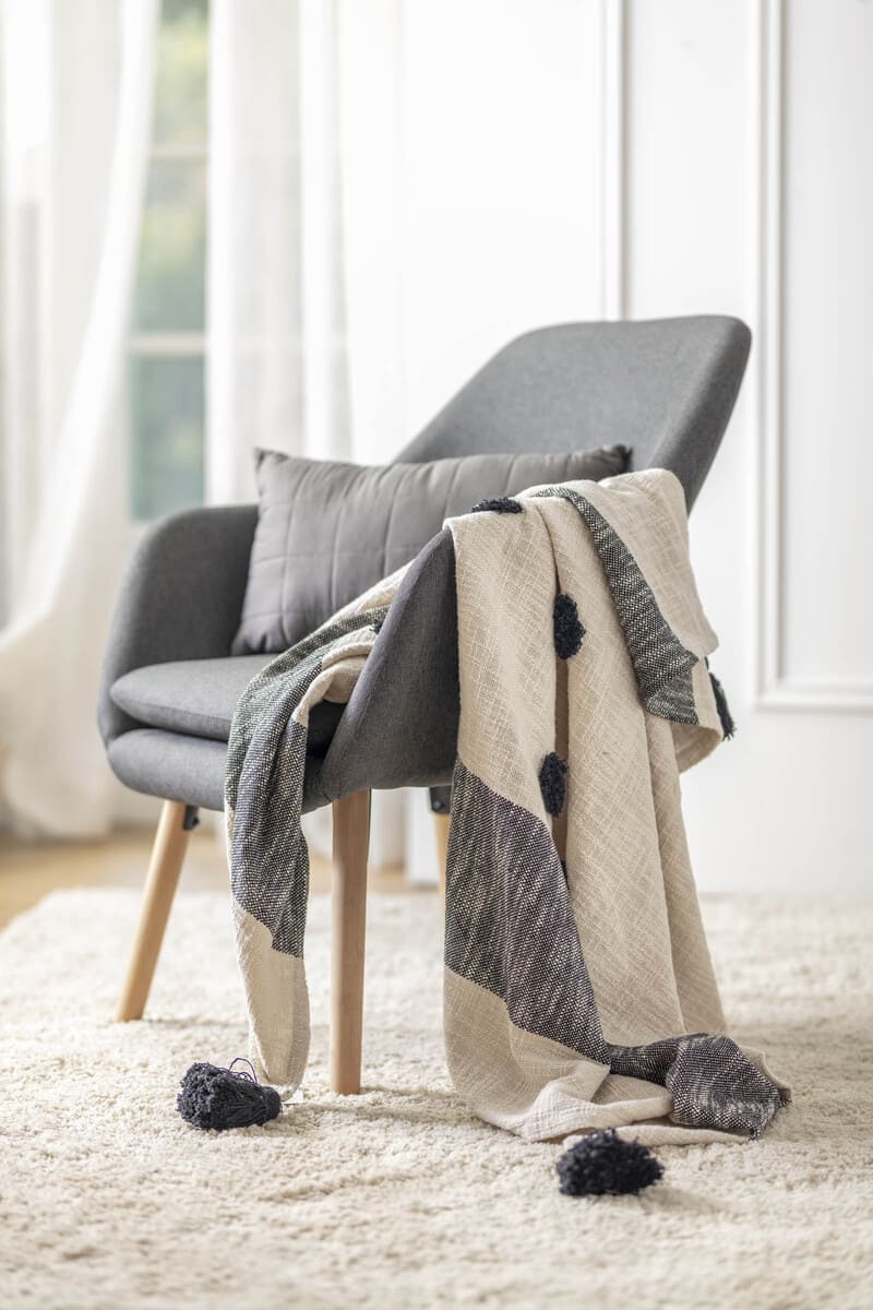 Poltrona gris patas de madera clara sobre una alfombra shaggy blanca. En la poltrona hay un cojín gris y una manta color crema y gris. Al fondo hay una ventana con cortinas blancas.