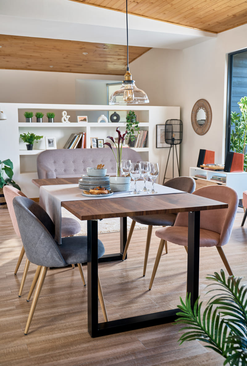 Living comedor con una mesa de madera y patas de metal negro rodeado con cuatro sillas de tela de distintos colores. Al fondo hay un sofá gris con respaldo estilo capitoné.